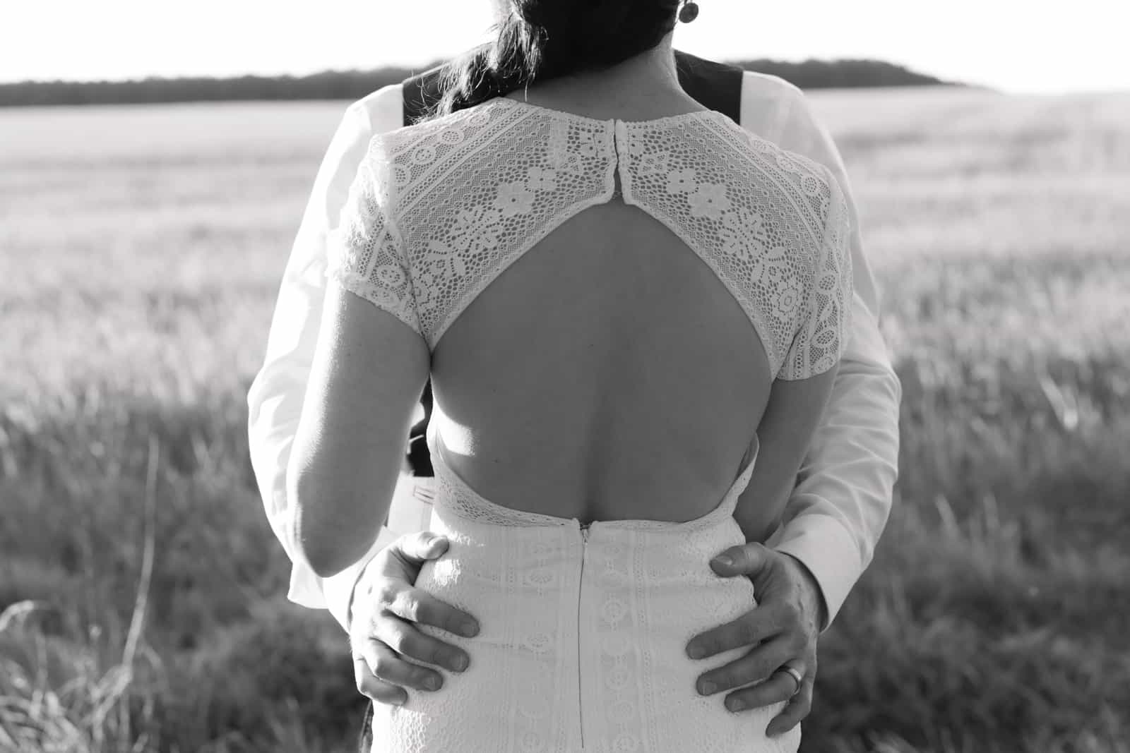 Brautpaar umarmt sich von hinten fotografiert in einem Kornfeld, Braut trägt Kleid mit Spitzendetails, Hochzeitsfotografie Brandenburg
