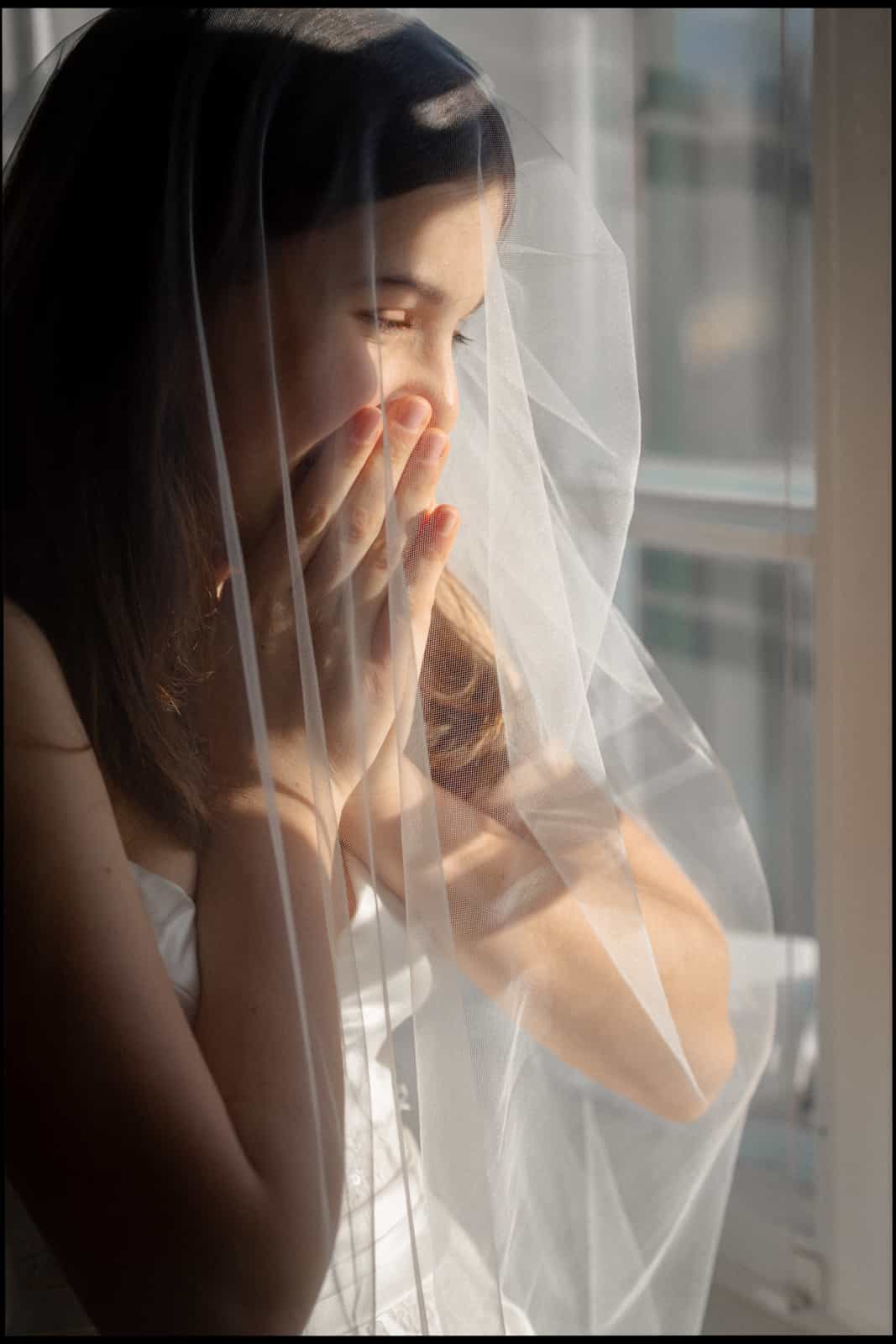 Bride with veil by window, hand covering mouth in emotional moment, natural light, documentary wedding photography Berlin