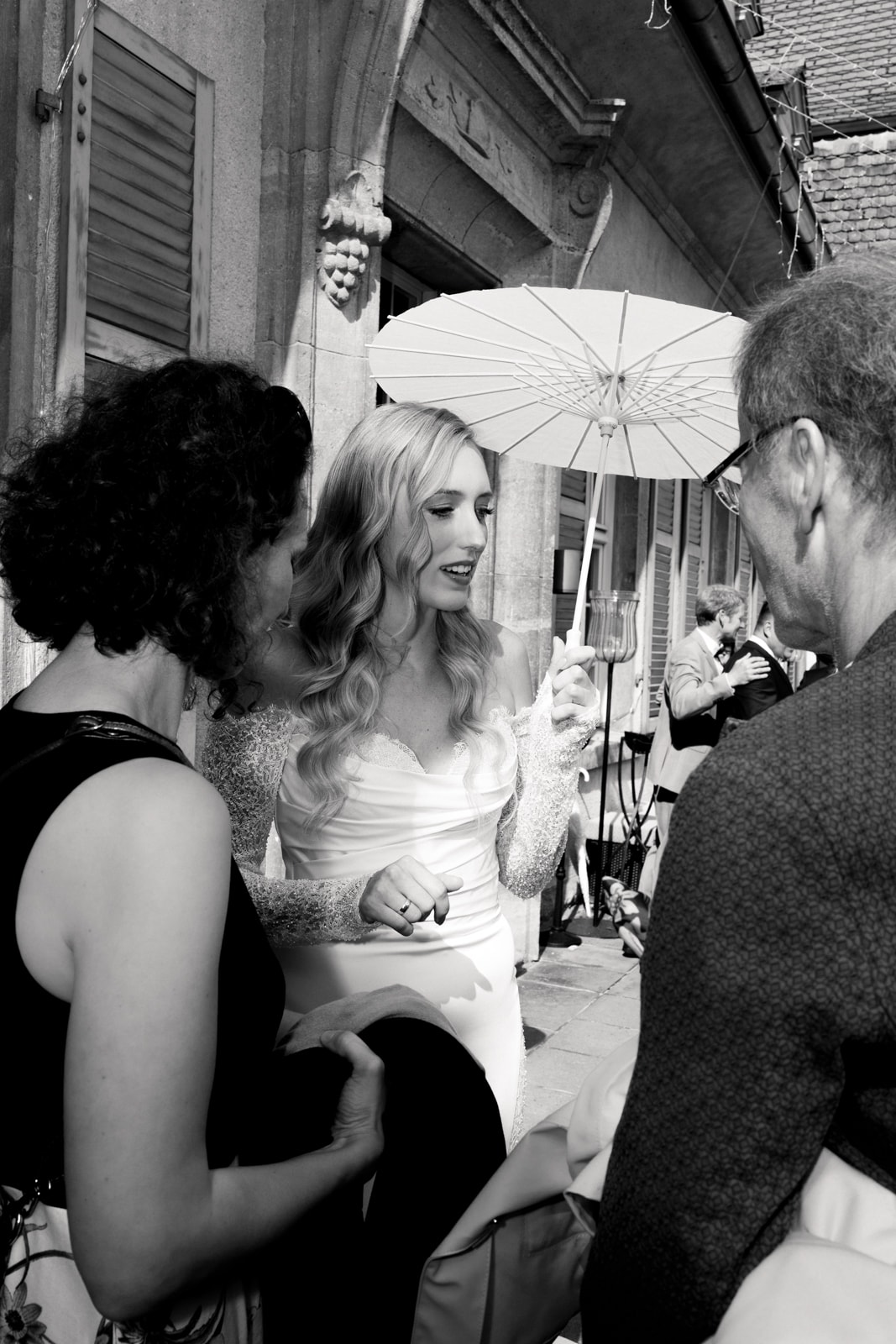 Blonde bride in elegant dress holding white parasol in front of historic building
