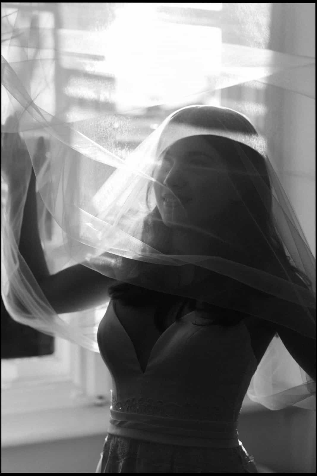 Bride from behind with flowing veil in window light, black and white documentary wedding photography