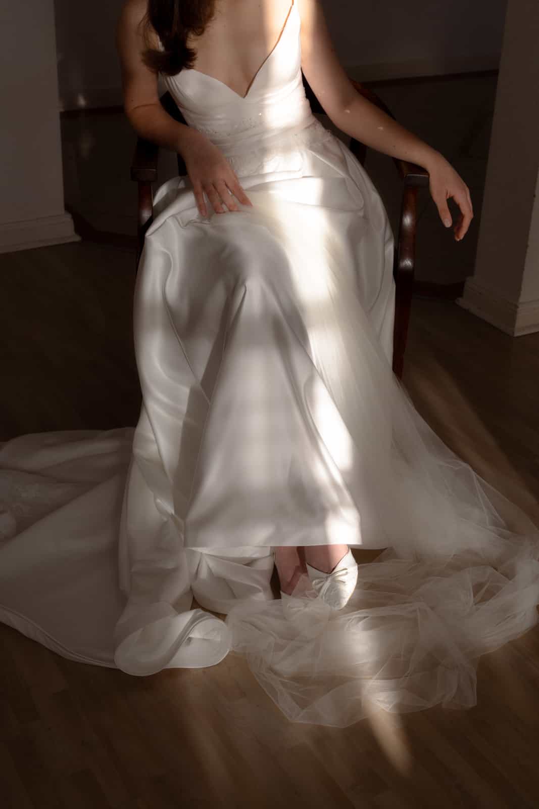 Bride in elegant white silk wedding dress sitting on antique chair surrounded by natural light and tulle