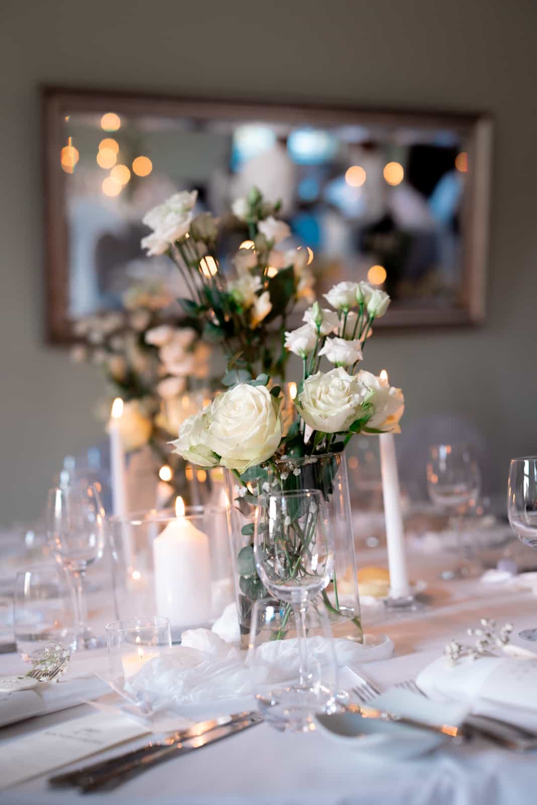 Elegantly set wedding table with white roses in glass vases, candles, and bokeh lights in the background