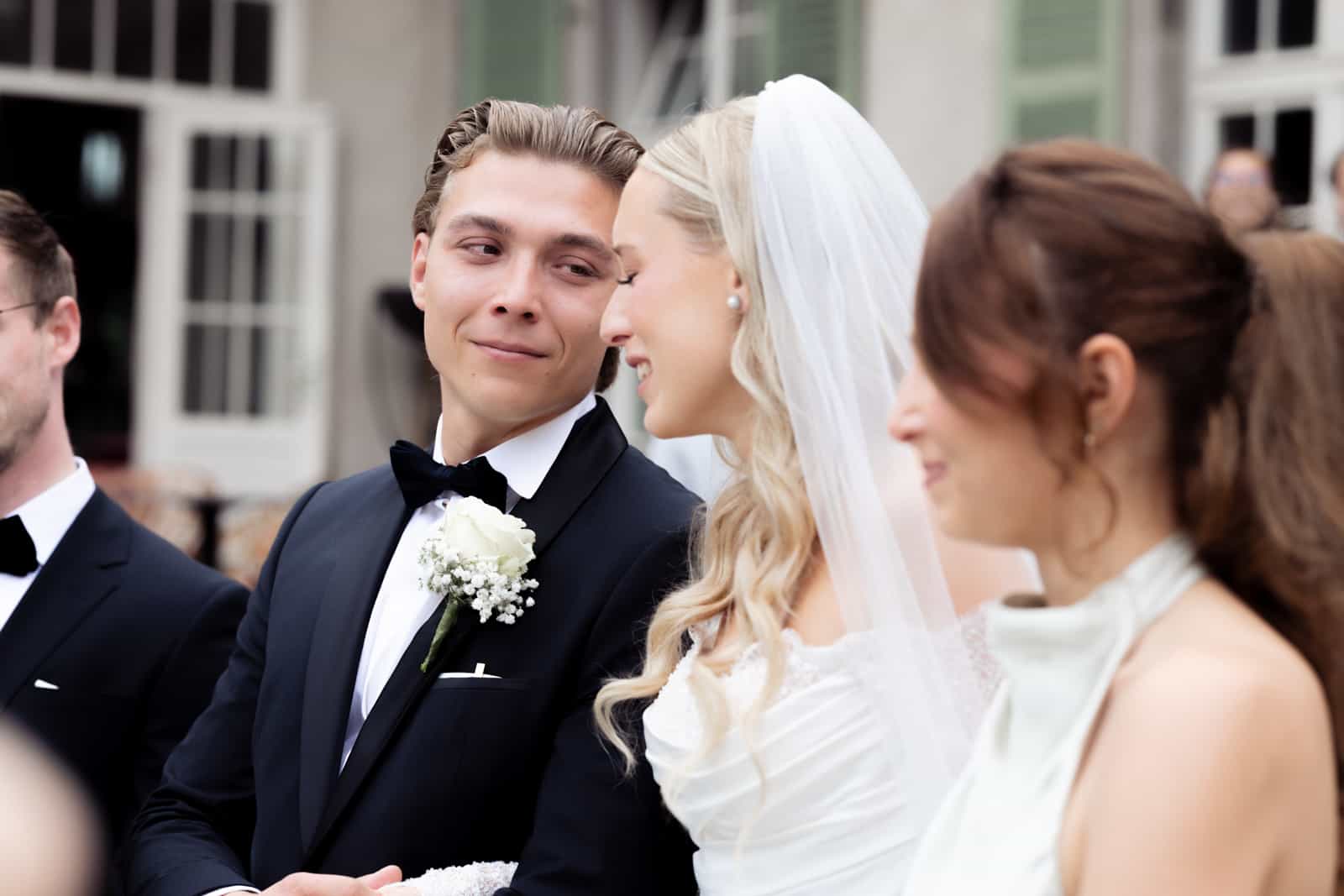 Groom in black tuxedo with bow tie and white boutonniere looking emotional during civil ceremony, bride with veil smiling beside him