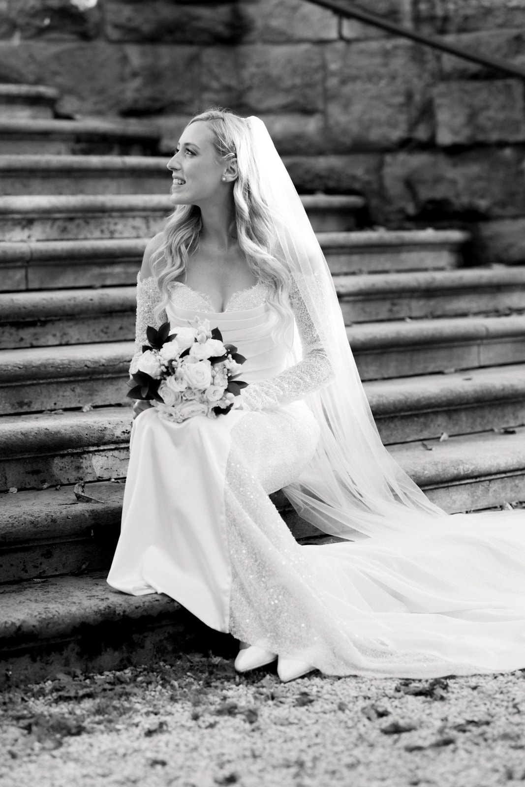 Bride in elegant lace dress with veil sitting radiantly on historic stone steps