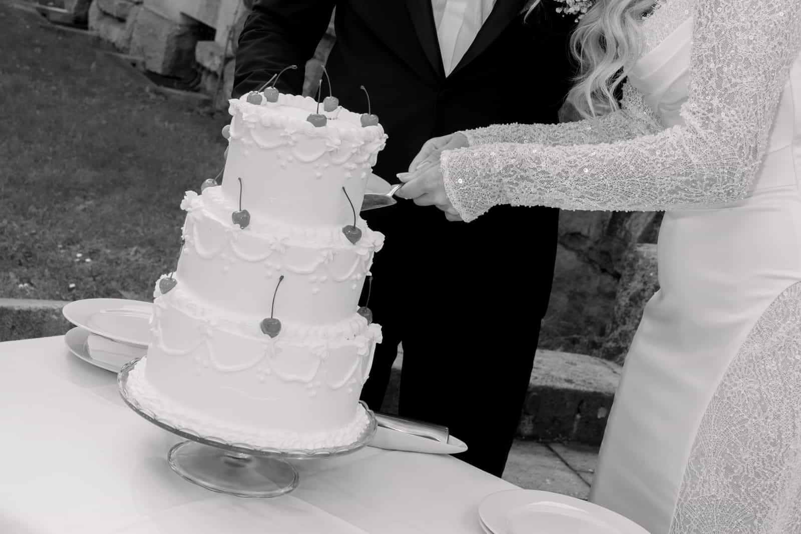 Bride and groom cutting multi-tiered white wedding cake with heart decorations together, bride in lace-detailed dress, groom in black suit