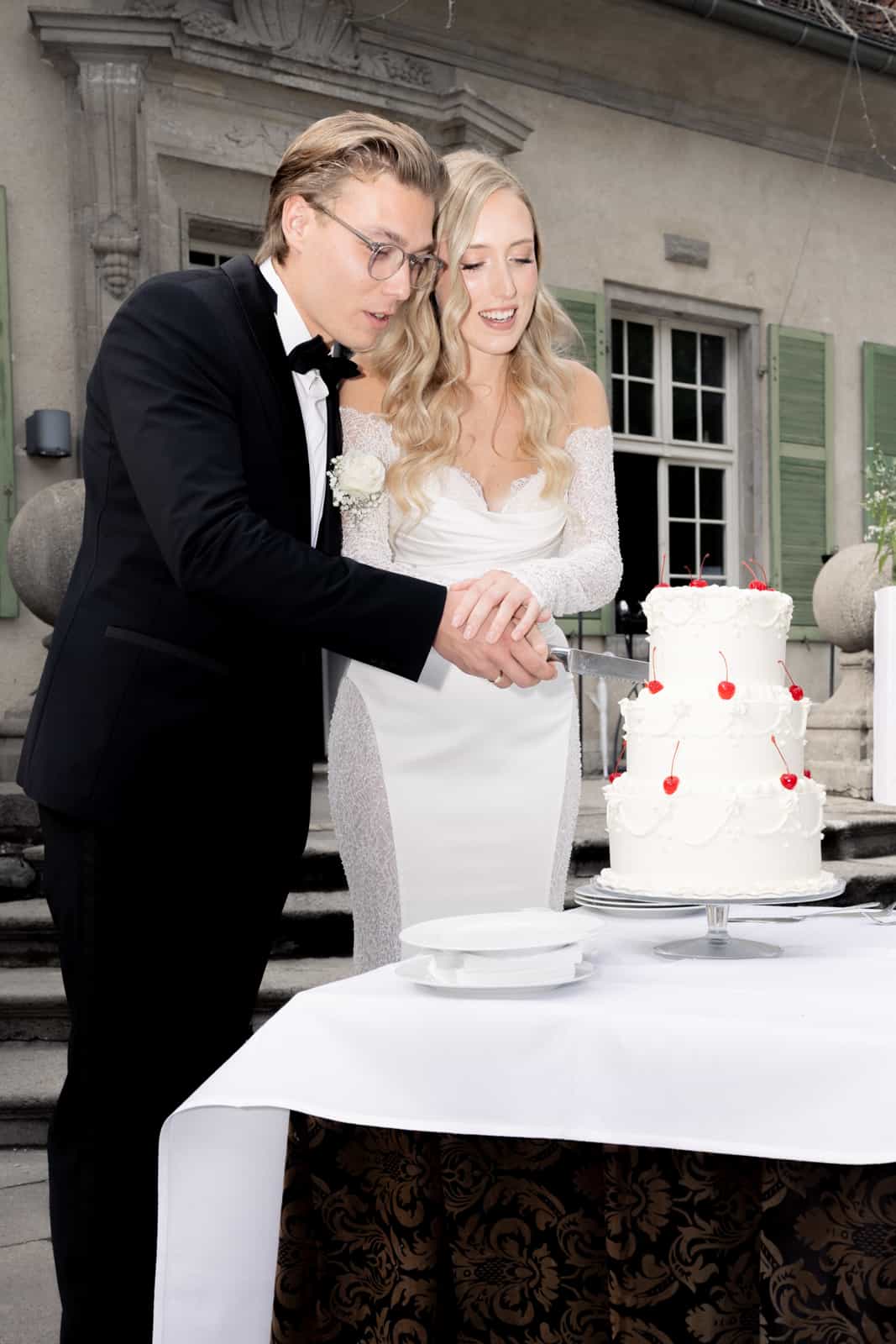 Bride and groom cutting white wedding cake decorated with red cherries together at historic venue