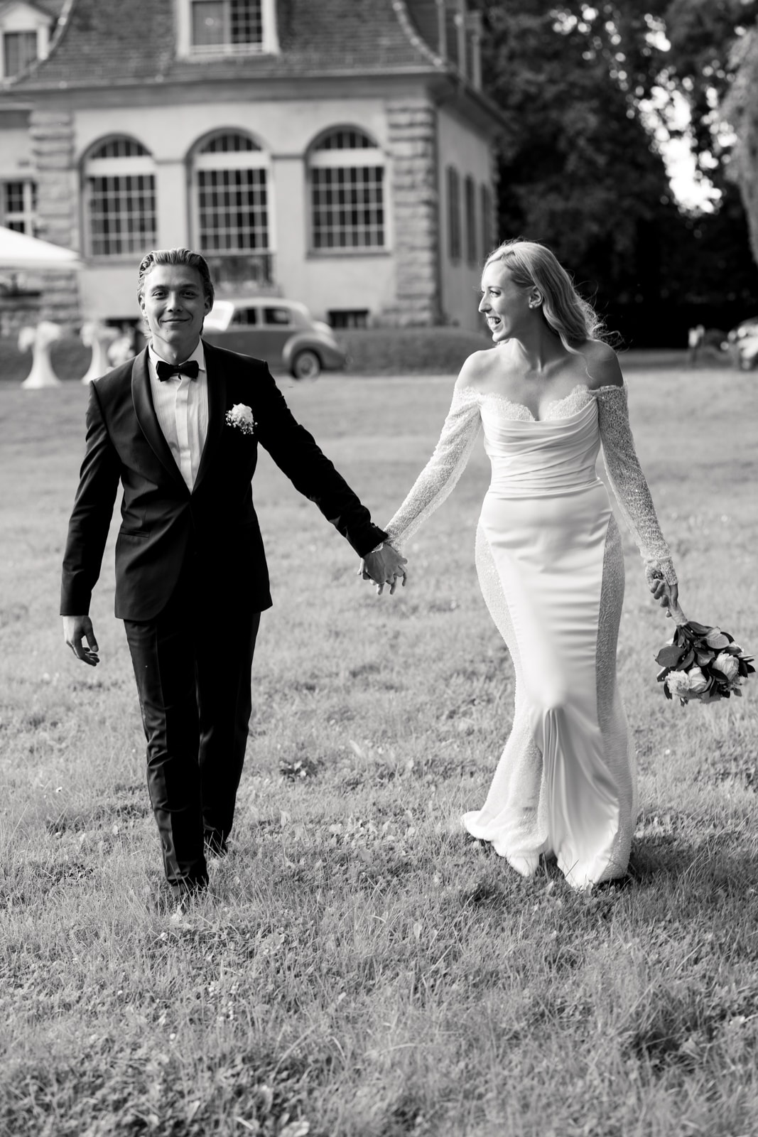 Wedding couple walking hand in hand across lawn in front of historic mansion building