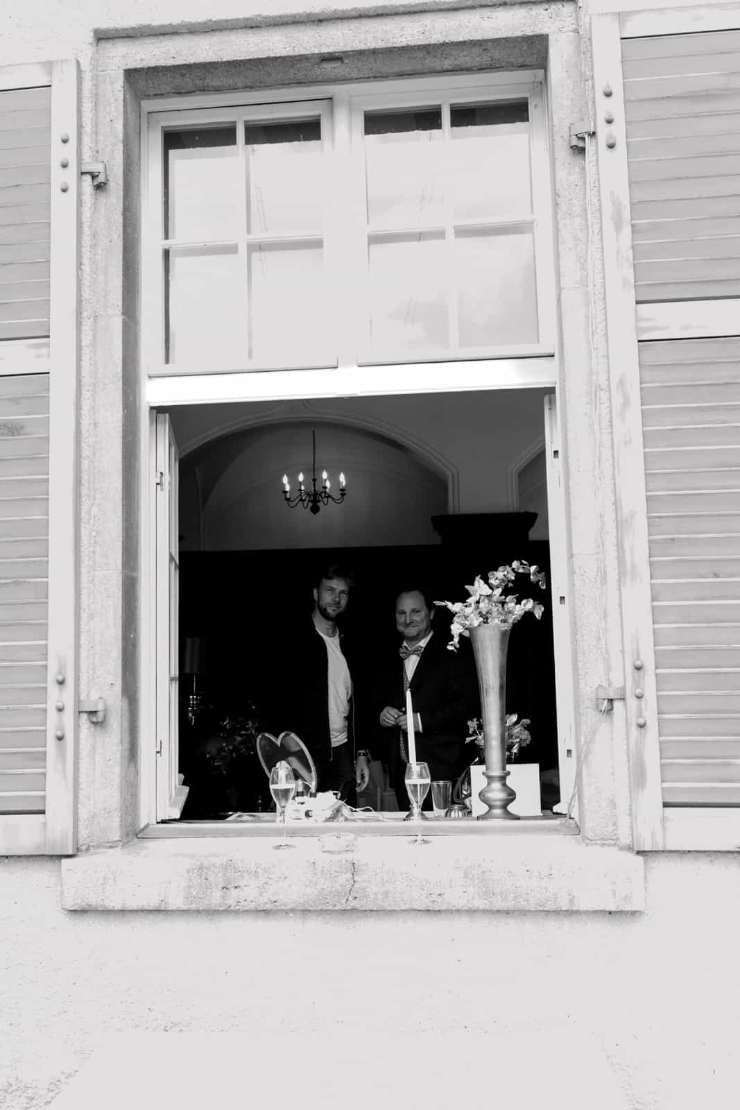 Two wedding guests in black suits standing at an open window of a wedding venue with chandelier and floral decoration, black and white photography
