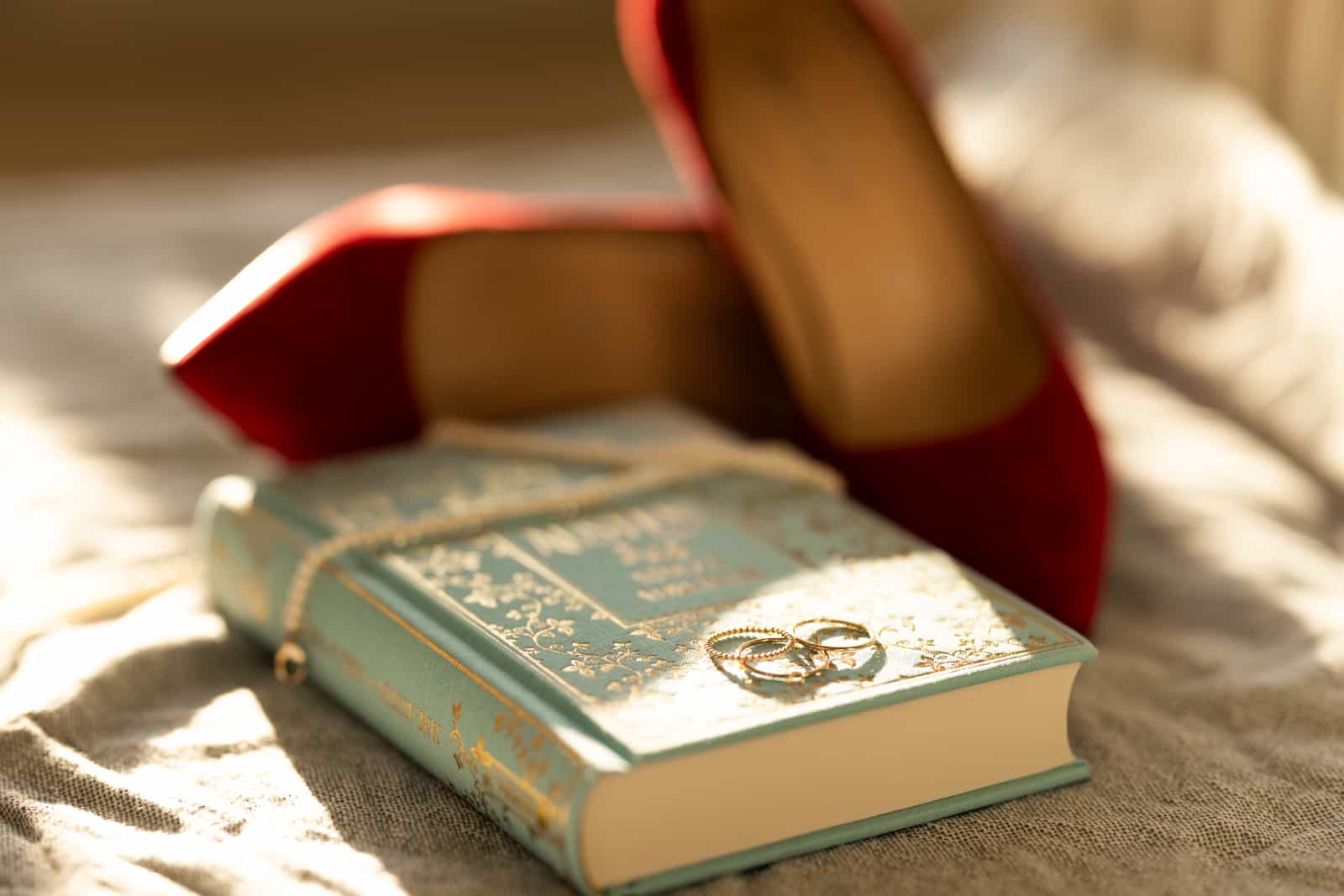 Golden wedding rings on antique turquoise book with red bridal shoes in background